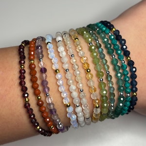May include: A selection of beaded bracelets in a range of colours, such as deep red, orange, purple, white, yellow, and green and blue hues. Each bracelet has small, faceted beads and gold or silver accents. The bracelets are stacked on a wrist.