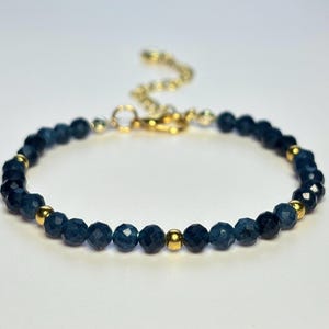 Puede incluir: Una delicada pulsera con cuentas facetadas de color azul oscuro intercaladas con pequeñas cuentas doradas. La pulsera tiene una cadena y un cierre dorados. La joya está sobre un fondo blanco.