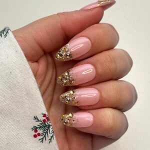 May include: Pink artificial nails with gold glitter and rhinestones at the tips. The nails are oval-shaped and have a glossy finish. The design features a gradient effect, transitioning from pink to gold. The nails are on a hand.