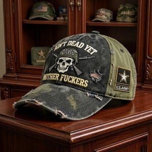 Puede incluir: Gorra de béisbol negra y verde oliva desgastada con el texto "Ain't Dead Yet Mother Fuckers" y un gráfico de calavera. Cuenta con un parche del ejército de EE. UU. y una estética vintage desgastada.