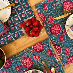 Peut inclure: Un ensemble de sets de table matelassés sur fond bleu sarcelle avec un motif répétitif de fleurs roses et de vignes vertes. Chaque set est accompagné d'une assiette blanche, de couverts dorés, d'une serviette assortie et d'un bol de fraises.