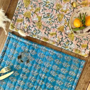 May include: Two floral placemats in pink and turquoise. The pink placemat features a large floral pattern with yellow and white accents. The turquoise placemat has a smaller, repeating floral design. A plate with oranges and a spoon are on the pink placemat.