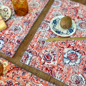 Pode incluir: Conjunto de individuais acolchoados retangulares com um padrão floral em coral, branco e azul. Os individuais apresentam um design repetido de flores estilizadas. Um copo de líquido laranja e um prato pequeno são visíveis.