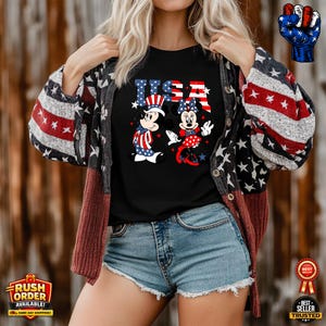 Puede incluir: Camiseta negra con la palabra "USA" en rojo, blanco y azul. Mickey y Minnie Mouse visten trajes patri&oacute;ticos. La persona lleva una rebeca con un estampado de estrellas y rayas y unos pantalones cortos vaqueros azules.
