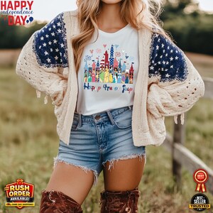 Puede incluir: Una camiseta blanca con un gr&aacute;fico de castillo colorido y el texto "Happy 4th of July". La camiseta se usa con pantalones cortos de mezclilla y una rebeca color crema con mangas con estampado de estrellas azules. La imagen tambi&eacute;n incluye el texto "Happy Independence Day."