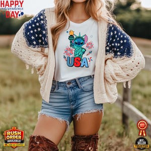Puede incluir: Camiseta blanca con un gr&aacute;fico de Stitch como la Estatua de la Libertad sosteniendo una antorcha, con la palabra "USA" en rojo, blanco y azul. La camiseta se usa con pantalones cortos de mezclilla y una rebeca color crema con mangas con estampado de estrellas azules. La imagen incluye el texto "Happy Independence Day".