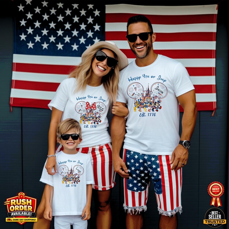 以下が含まれることがあります： ミッキーマウスの耳のグラフィックと「Happy 4th of July Est. 1776」の文字が入ったお揃いの白いTシャツを着た家族。父親は星とストライプのデザインのショーツを着用しています。背景にはアメリカ国旗があります。