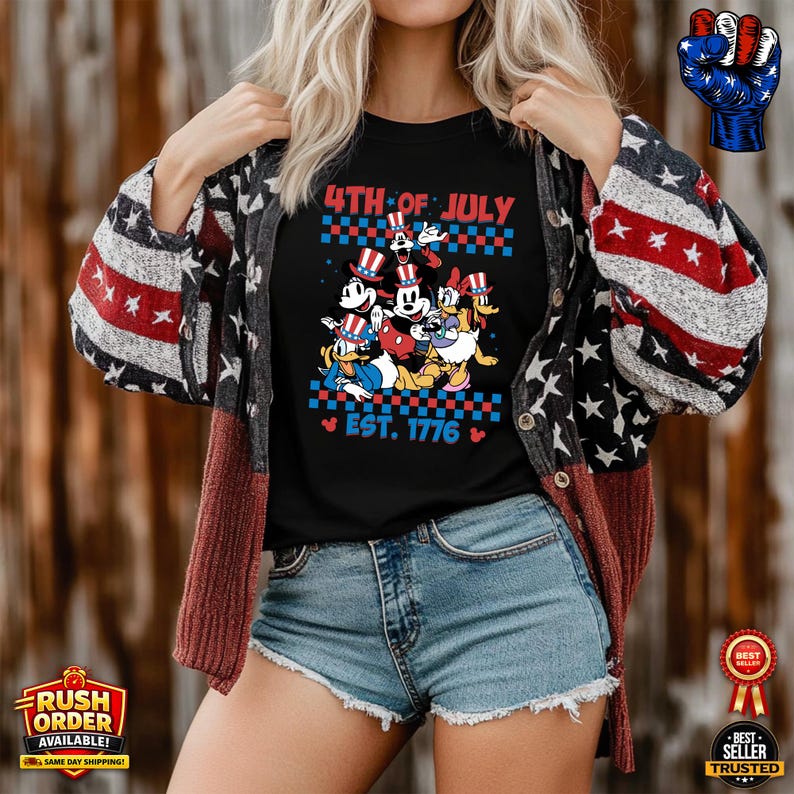 Puede incluir: Camiseta negra con un dise&ntilde;o de Mickey, Minnie, Donald y Goofy con atuendos patri&oacute;ticos. La camiseta dice "4th of July" y "Est. 1776". La persona lleva una rebeca con un estampado de estrellas y rayas.