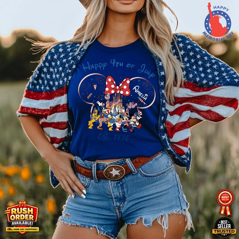 以下が含まれることがあります： ミッキーマウスの耳、赤いリボン、そして「Happy 4th of July Amelia Est. 1776」の文字が特徴的な愛国的なデザインの青いTシャツ。その人は、赤、白、青のアメリカ国旗のデザインのジャケットを着ています。