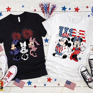 Puede incluir: Dos camisetas, una negra y otra blanca, con dise&ntilde;os patri&oacute;ticos. La camiseta negra tiene a Minnie Mouse y fuegos artificiales. La camiseta blanca muestra a Mickey y Minnie Mouse con el texto "USA". Banderas americanas y estrellas decoran el conjunto.
