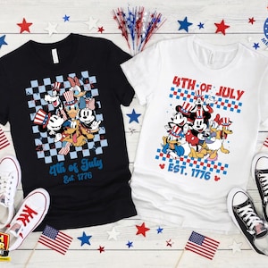 Puede incluir: Dos camisetas, una negra y otra blanca, con personajes de dibujos animados con atuendos patri&oacute;ticos. Las camisetas muestran "4th of July Est. 1776". Banderas americanas y estrellas decoran las camisetas, junto con pares de zapatillas.