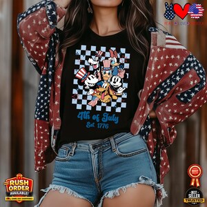Puede incluir: Camiseta negra con un dise&ntilde;o de personajes de Disney celebrando el 4 de julio. El dise&ntilde;o incluye a Mickey Mouse, el Pato Donald y Goofy, sobre un fondo a cuadros. El texto "4th of July Est. 1776" se muestra en azul.