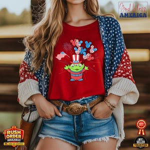 Puede incluir: Camiseta roja con un alien&iacute;gena de dibujos animados con un sombrero de copa patri&oacute;tico y orejas de Mickey Mouse. El dise&ntilde;o incluye fuegos artificiales y una peque&ntilde;a bandera estadounidense. La camiseta se combina con pantalones cortos de mezclilla y una rebeca con un patr&oacute;n de estrellas y rayas.
