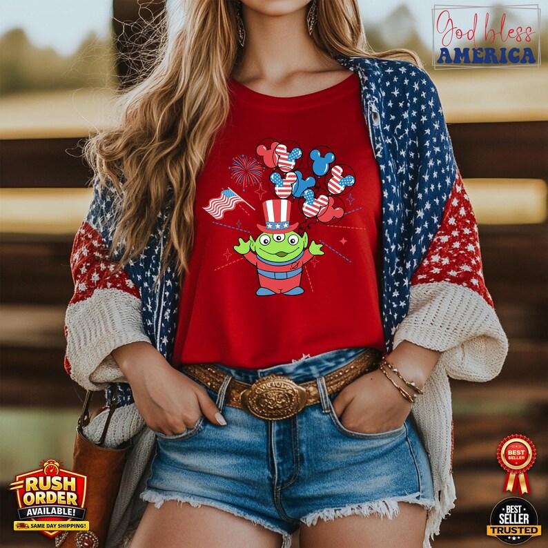 Puede incluir: Camiseta roja con un alien&iacute;gena de dibujos animados con un sombrero de copa patri&oacute;tico y orejas de Mickey Mouse. El dise&ntilde;o incluye fuegos artificiales y una peque&ntilde;a bandera estadounidense. La camiseta se combina con pantalones cortos de mezclilla y una rebeca con un patr&oacute;n de estrellas y rayas.