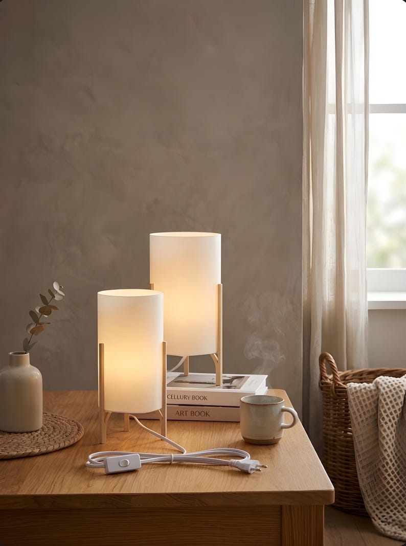 May include: Two white cylindrical table lamps with wooden frames on a wooden table. The lamps are lit, and a white power cord with a switch is visible. A vase, books, and a mug are also on the table.