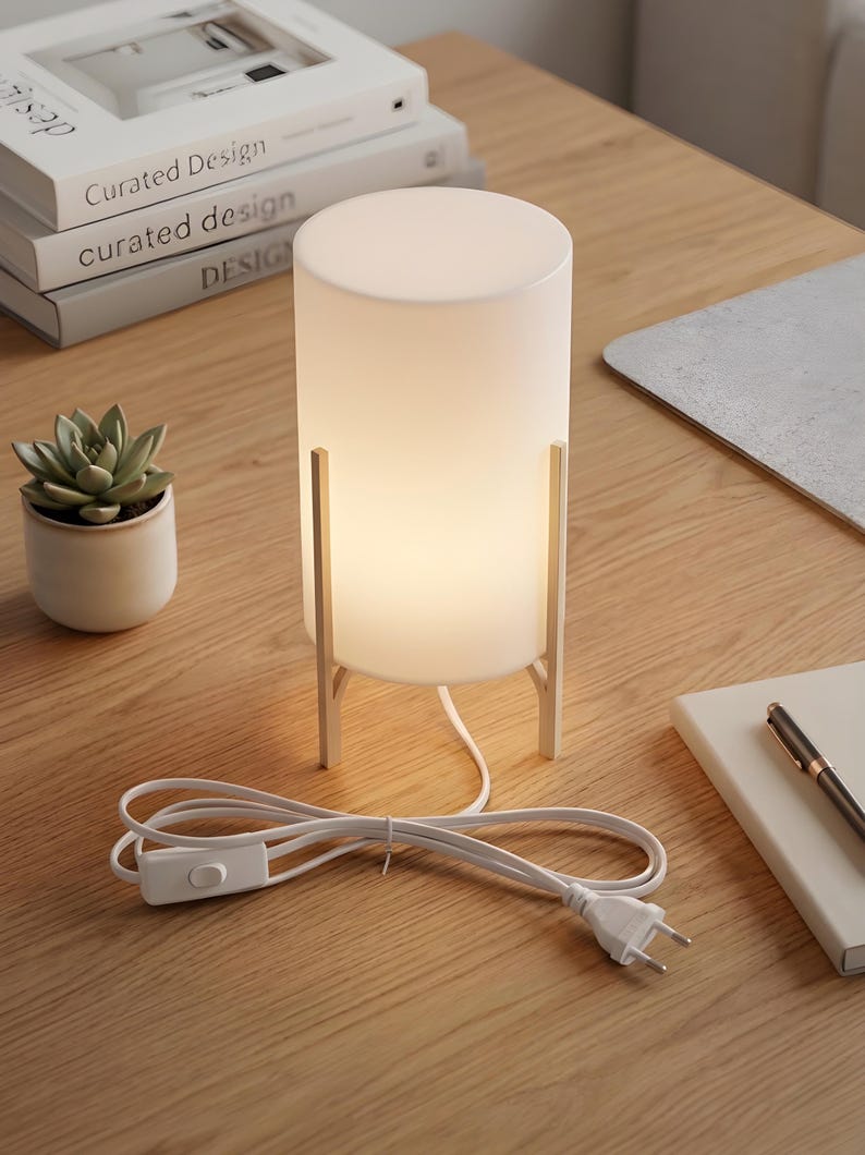 May include: A contemporary white cylindrical table lamp with a soft glow, supported by a silver metal frame. The lamp is on a wooden surface, with a succulent plant in a small pot and books nearby.