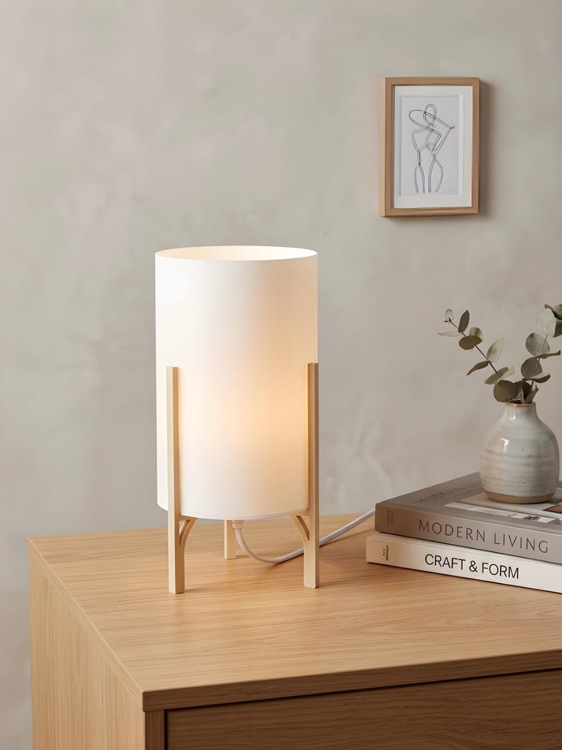 May include: A cylindrical white table lamp with a warm glow, supported by a light wood frame. The lamp is on a wooden surface, next to books and a small vase. A framed art print hangs on the wall.