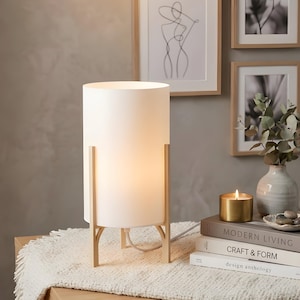 May include: A white cylindrical table lamp with a warm glow, supported by a light wood frame. The lamp sits on a wooden bedside table with a fringed white runner. Framed art and a candle are in the background.