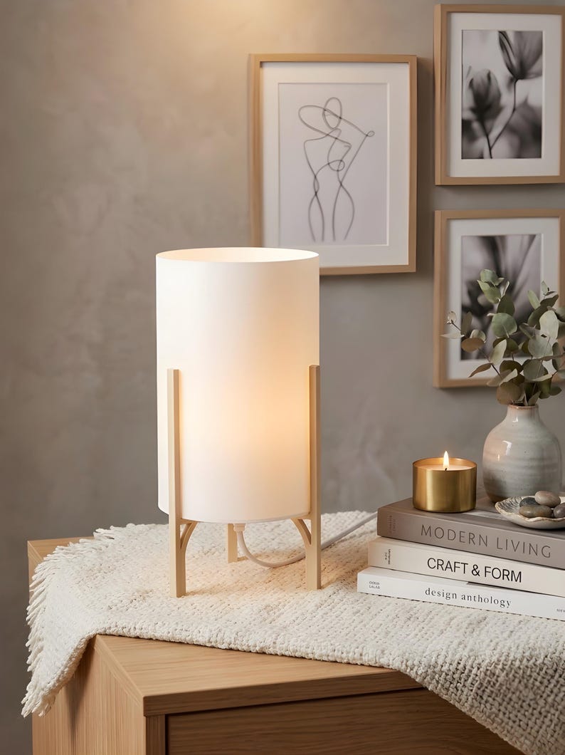 May include: A white cylindrical table lamp with a warm glow, supported by a light wood frame. The lamp sits on a wooden bedside table with a fringed white runner. Framed art and a candle are in the background.