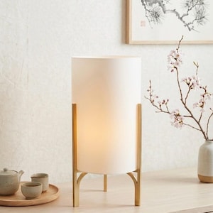 May include: A white cylindrical lamp with a gold-coloured metal frame. The lamp is illuminated, emitting a warm light. It sits on a light wood surface, with a tea set and framed artwork in the background. The style is minimalist and modern.