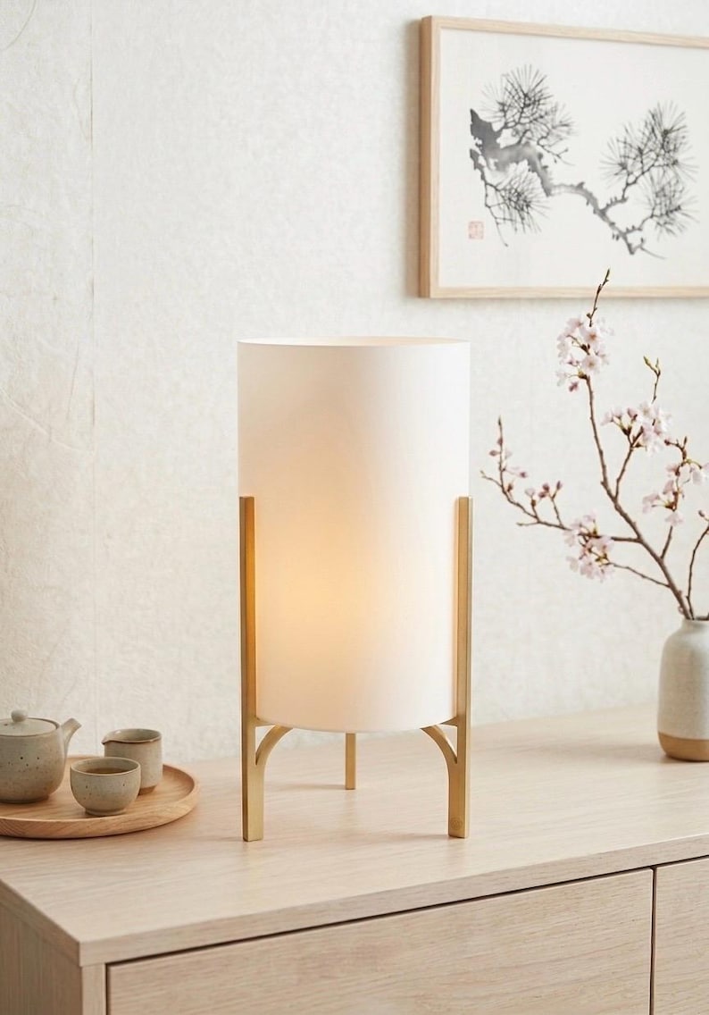 May include: A white cylindrical lamp with a gold-coloured metal frame. The lamp is illuminated, emitting a warm light. It sits on a light wood surface, with a tea set and framed artwork in the background. The style is minimalist and modern.