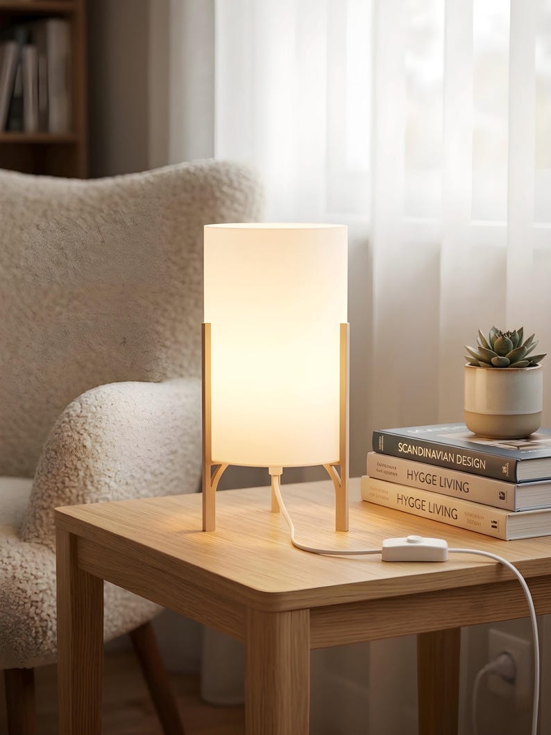 May include: A contemporary table lamp featuring a white cylindrical shade supported by a light wood frame. It sits on a wooden side table, alongside a small potted succulent and books. The lamp is plugged in.