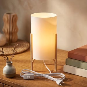May include: A white cylindrical table lamp with a warm glow, supported by a light wood frame. The lamp has a white cord with an on/off switch and a plug. The lamp is on a wooden surface.
