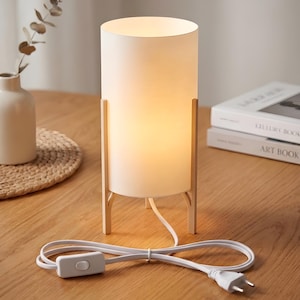 May include: A white cylindrical table lamp with a warm glow, supported by a light wood frame. The lamp has a white cord with an on/off switch and a two-pin plug. The lamp is on a wooden table.