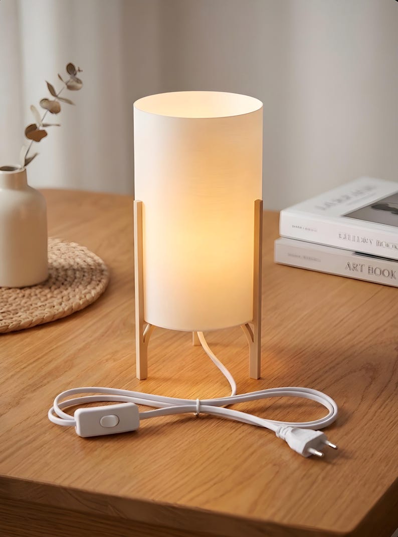 May include: A white cylindrical table lamp with a warm glow, supported by a light wood frame. The lamp has a white cord with an on/off switch and a two-pin plug. The lamp is on a wooden table.