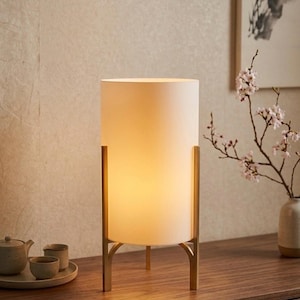 May include: A cream-coloured cylindrical table lamp with a warm glow, supported by a gold-coloured metal frame. The lamp sits on a wooden surface, with a tea set and a vase of flowers in the background. The lamp's height is approximately 30 cm.
