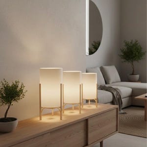 May include: Three contemporary table lamps with cylindrical white shades and gold-coloured frames are arranged on a wooden cabinet. The lamps are evenly spaced, lighting a living room with a neutral colour scheme.