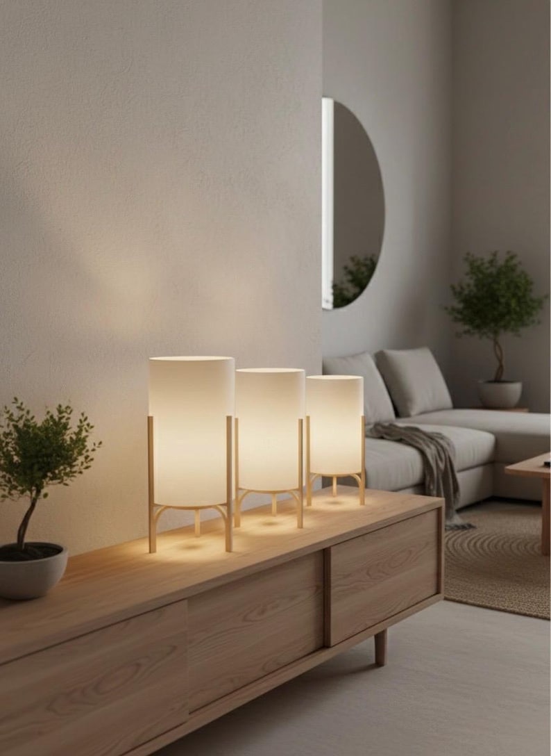 May include: Three contemporary table lamps with cylindrical white shades and gold-coloured frames are arranged on a wooden cabinet. The lamps are evenly spaced, lighting a living room with a neutral colour scheme.