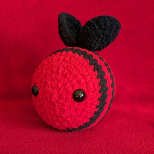 May include: A crocheted bee plush toy with black and red stripes and black eyes. The bee has two black crocheted wings.