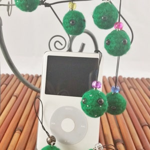 Puede incluir: Un conjunto de nueve adornos de bolas de fieltro verde con ojos negros y cuentas de varios colores. Los adornos cuelgan de un marco de alambre negro y un iPod blanco.