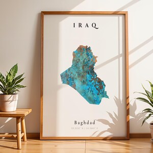 May include: A framed print depicting a map of Iraq in shades of blue and brown. The word "IRAQ" is printed at the top, and "Baghdad" is at the bottom. The print is displayed in a room with plants and a small wooden stool.