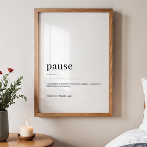 May include: A framed print featuring the word "pause" and its definition. The print is in a wooden frame, hanging on a neutral wall. The definition describes a small break that restores more than it takes.