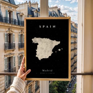 以下が含まれることがあります： 黒い背景に、明るい色のスペインの地図が描かれた額入りのプリント。上部には「SPAIN」、下部には座標とともに「Madrid」の文字。フレームは金色です。