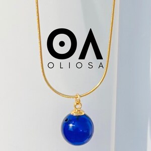 Peut inclure: Un collier à chaîne dorée avec un pendentif sphérique bleu foncé. Le pendentif est suspendu à une petite bélière dorée. Le collier est sur fond blanc, avec le mot "OLIOSA" en lettres noires.