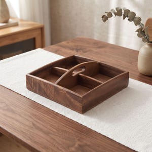 May include: A square wooden serving tray with a dark brown finish, featuring four distinct compartments separated by intersecting dividers and a central arched handle with a cutout grip for easy carrying and organization of small items.