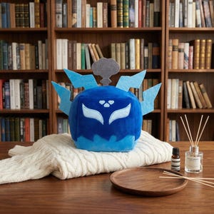 May include: A plush blue character with light blue accents and a gray cloud detail. The character is sitting on a cream-colored sweater, with a wooden tray, essential oil bottle, and diffuser in the foreground. Bookshelves fill the background.