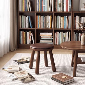 Può includere: Uno sgabello rotondo in legno con seduta imbottita marrone scuro e quattro gambe robuste. Lo sgabello è posizionato su un tappeto bianco testurizzato, circondato da diversi libri con copertina rigida sparsi sul pavimento vicino a un tavolino e una libreria.