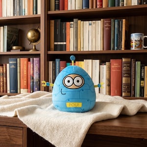 May include: A blue plush robot toy with big eyes, a small antenna, and yellow arms. The robot has the word "POU!" on its front. The toy is on a white blanket in front of a bookshelf filled with books.