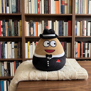 Może przedstawiać: Pluszowa zabawka w kształcie ziemniaka, ubrana w czarny smoking, muszkę i mały czarny kapelusz. Zabawka ma duże, kreskówkowe oczy i czerwone, uśmiechnięte usta. Siedzi na kremowym, dzianinowym kocu przed półką z książkami.