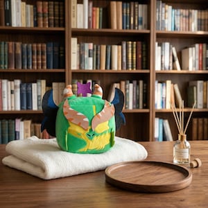 Könnte beinhalten: Ein grüner Plüschdrache mit orangefarbenen und weißen Akzenten, schwarzen Flügeln und einem lila Stern. Er liegt auf einer gefalteten weißen Decke, neben einem Holztablett, einem Diffusor und einem Bücherregal im Hintergrund.