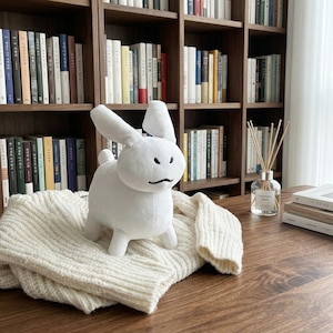 Puede incluir: Un juguete de peluche blanco con forma de conejito de diseño minimalista, con una pequeña cola y una cara sencilla, está sobre un suéter de punto color crema. El fondo incluye una estantería de madera llena de libros, creando una estética acogedora.
