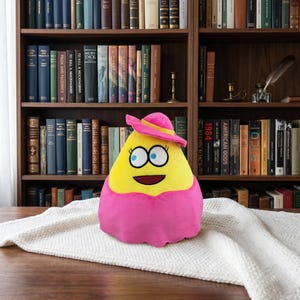 May include: A plush toy shaped like a yellow triangle with a pink hat and collar. The toy has large blue eyes, a smiling mouth, and is set against a backdrop of a bookshelf filled with books. The toy is sitting on a white, textured surface.