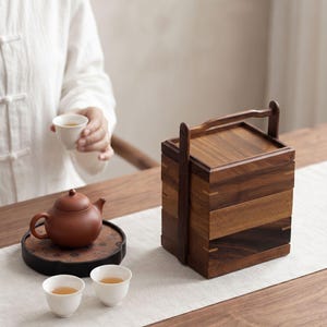 May include: A three-tiered wooden food carrier with a top handle, a brown clay teapot on a circular tray, and three small white ceramic tea cups. The carrier features dark wood grain panels with light wood joinery details on the corners.