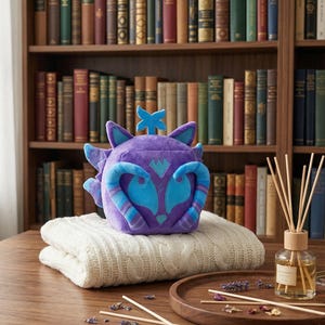Op de afbeelding: Een paars pluche dier met blauwe accenten en een blauwe ster zit op een gevouwen, crèmekleurige deken. Een glazen diffuser met houten stokjes en een houten dienblad met gedroogde bloemen zijn ook aanwezig.