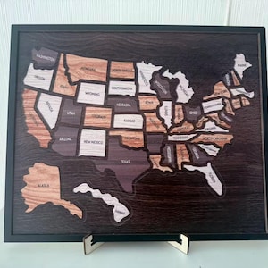 May include: A framed wooden map of the United States featuring individual state cutouts in varying shades of light, medium, and dark wood grain. Each state is labeled with its name, and the entire map is set against a dark wood background.