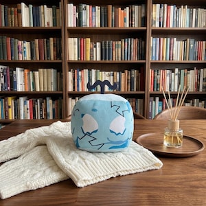 May include: A light blue cube-shaped plush toy with a dark blue mustache on top. The toy has a white and blue design. It sits on a white cable-knit sweater on a wooden table. A wooden tray with a glass bottle and reeds is also on the table.